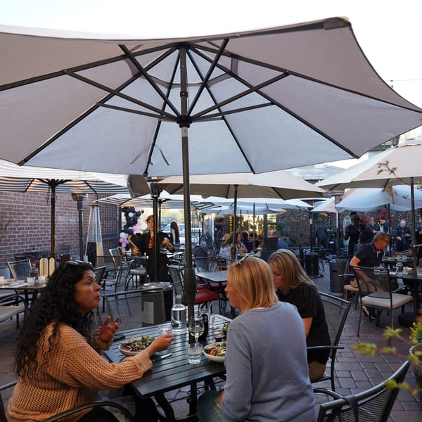 Patrons dining outdoors under large umbrellas at a restaurant patio with brick walls and string lights.