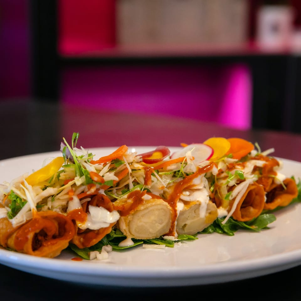 Crispy taquitos topped with fresh cabbage, carrots, radishes, crema, and red sauce on a white plate.