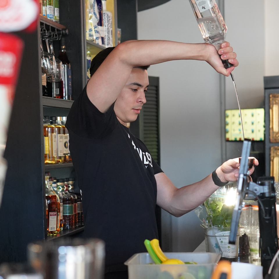 A bartender at La Condesa Eatery pours a drink, with bottles of liquor visible in the background.