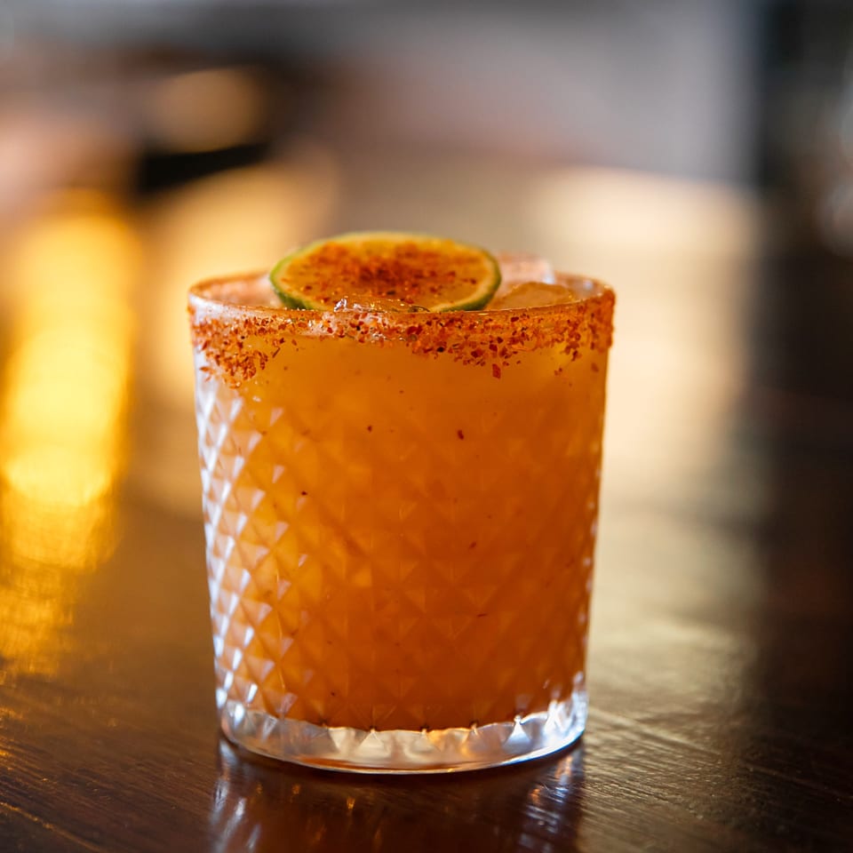 La Condesa Eatery's orange cocktail with a chili-lime rim and lime slice on a dark wooden table.