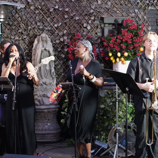 A band performs live with two female vocalists and a male trombonist on an outdoor patio.