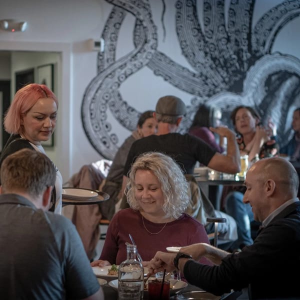 A La Condesa Eatery server with pink hair carries plates to a table of smiling diners.