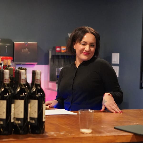 A woman winks behind a bar with wine bottles and a glass on a wooden counter.