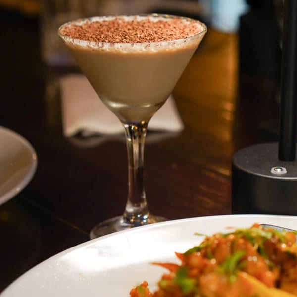 A chocolate martini with a cocoa-dusted rim sits on a dark table next to a plate of food.