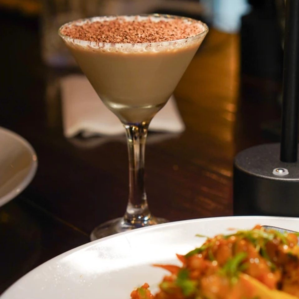 A chocolate martini with a cocoa-dusted rim sits on a dark table next to a plate of food.