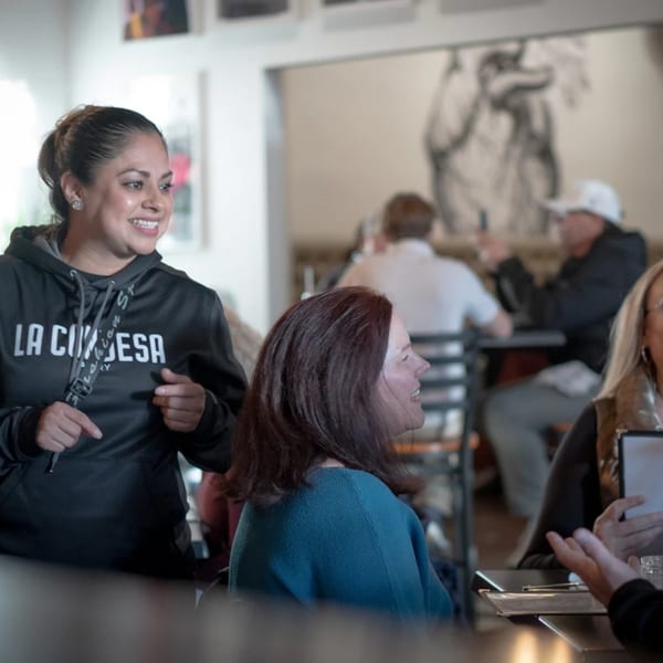 A smiling La Condesa Eatery staff member in a black hoodie talks to seated customers.