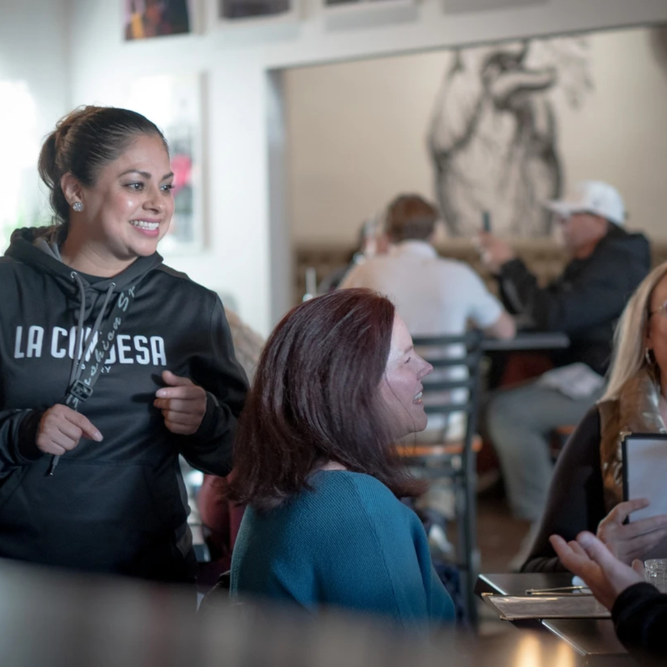 A smiling La Condesa Eatery staff member in a black hoodie talks to seated customers.