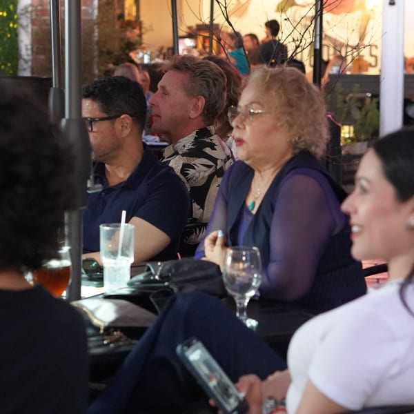 People dining at an outdoor restaurant, with drinks on the table and warm lighting.
