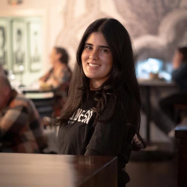 A smiling woman with long dark hair stands in a dimly lit restaurant, wearing a black t-shirt.