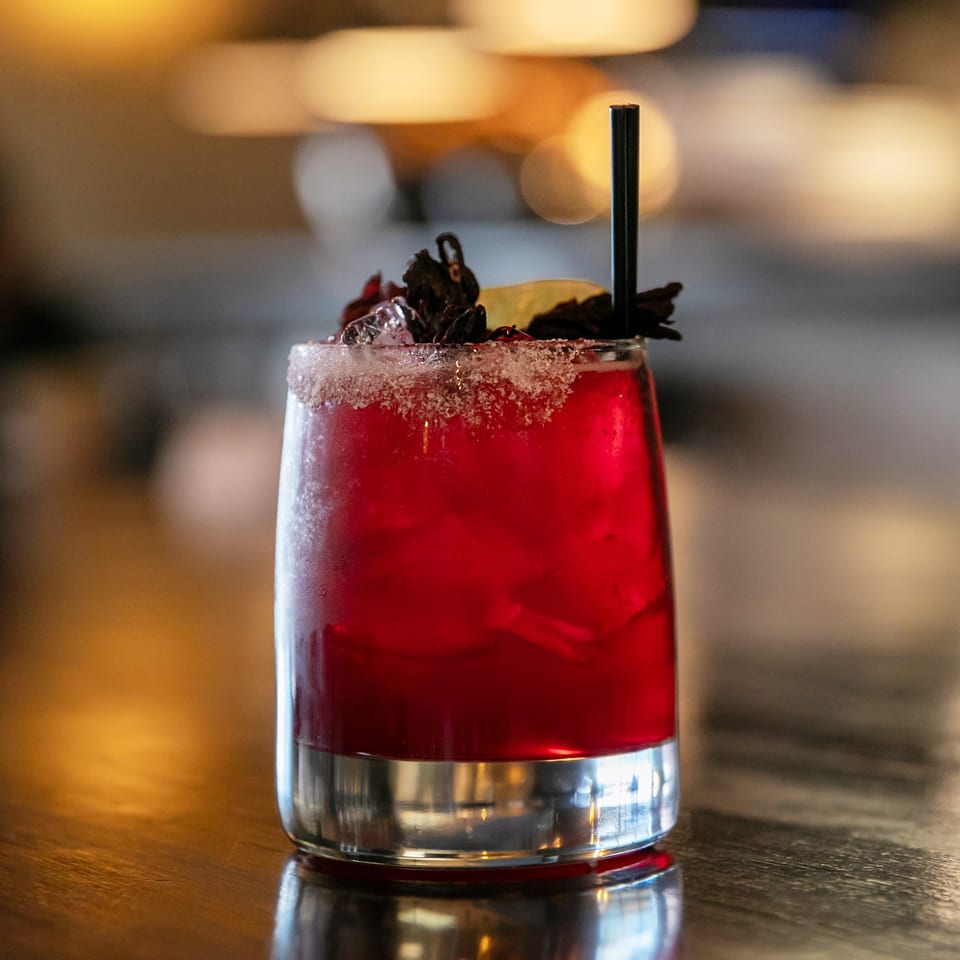 A vibrant red cocktail with a sugared rim, garnished with dried hibiscus and a lime slice.