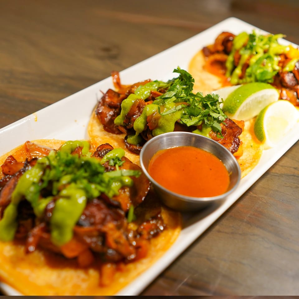 Three La Condesa Eatery fusion tacos with green sauce, cilantro, and lime wedges on a white plate.