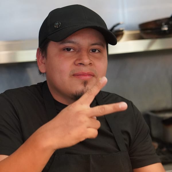 A La Condesa Eatery chef in a black cap and apron, making a peace sign.