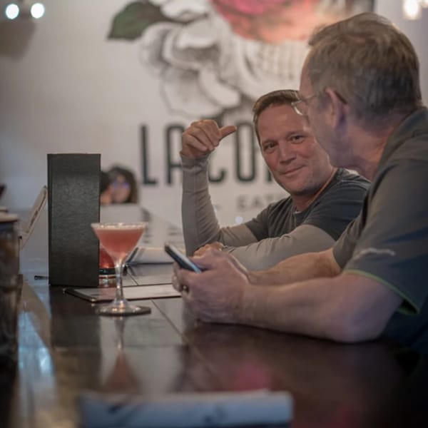 Two men conversing at a bar, with a pink cocktail and phone on the counter.