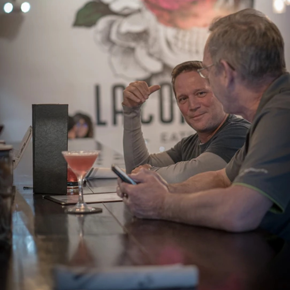 Two men conversing at a bar, with a pink cocktail and phone on the counter.