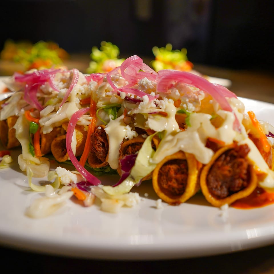 Crispy taquitos topped with shredded cabbage, pickled red onions, crumbled cheese, and crema.