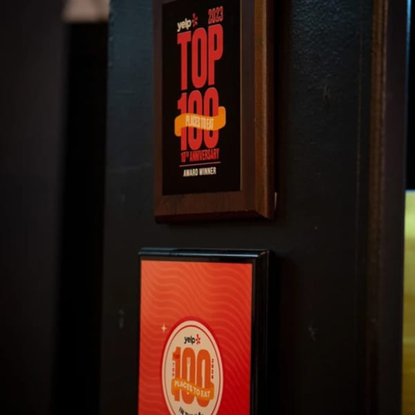Two framed Yelp "Top 100 Places to Eat" awards, one for 2023, displayed on a dark wall.