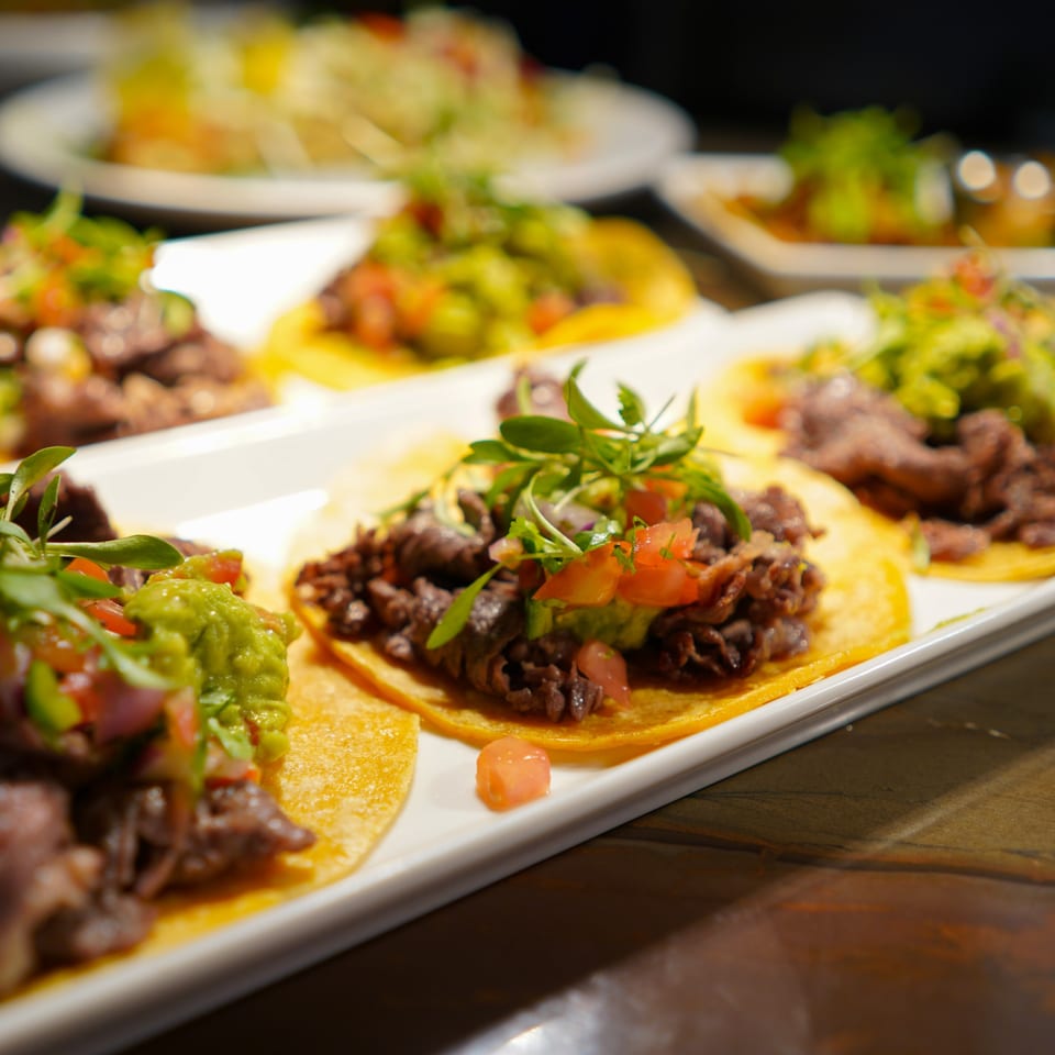 A white platter holds several La Condesa Eatery tacos with meat, guacamole, and microgreens.